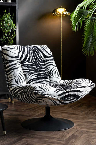 The Zebra Print Velvet Swivel Chair styled in front of a dark wall with a floor lamp and some plants.