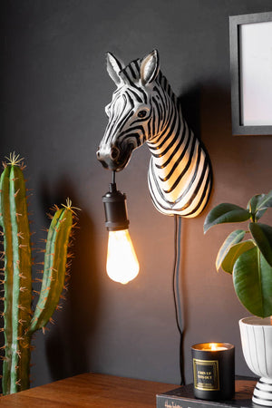 The Zebra Wall Light displayed on a black wall with some artwork, above various plants/home accessories.
