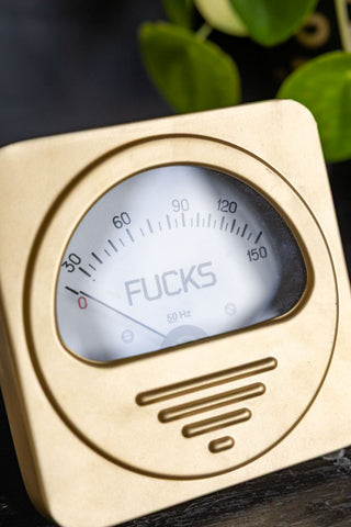 Close-up of the Zero Fucks Gold Ornament displayed on a dark wooden surface with some greenery in the background.