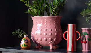 Lifestyle image of the Orange Hand-painted Floral Glass Vase, Extra Large Pink Footed Planter, Tall Red Stripe Vase With Handles and Pink Cherry Vase styled together on a black sideboard in front of a black wall. 