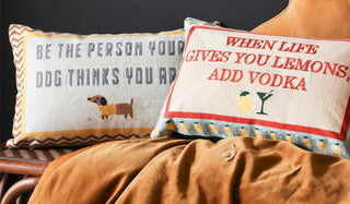 Lifestyle image of the Be The Person Your Dog Thinks You Are Cushion and the When Life Gives You Lemons Add Vodka Cushion styled together with other soft furnishings on a wicker bench in front of a black wall. 