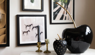 The Small Black & Gold Strawberry Ornament and Large Cherry Ornament styled together on a marble table with some gold candlesticks, with art prints on the wall in the background.