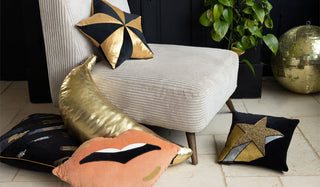A selection of new Rockett St George cushions displayed on/around a beige chair, with a black sideboard, plant and gold disco ball in the backlground.