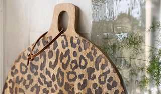 Lifestyle image of the Lifestyle image of the Leopard Print Mango Wood Serving Board displayed in front of a tiled kitchen wall and styled with a plant. 