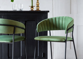 Lifestyle image of two of the Curved Back Velvet Bar Stool In Moss Green Bar Stools styled in front of a black cabinet with decorative accessories on the top. 