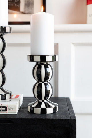 A black and white striped candle holder with a lit pillar candle inside, styled on a black sideboard with a book and another candle holder.