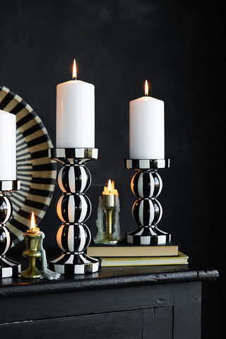 Two monochrome striped candle holders with lit candles inside, styled on a black sideboard with some other home accessories.