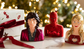 Three quirky celebrity-inspired baubles styled on a white surface with a wrapped Christmas present.