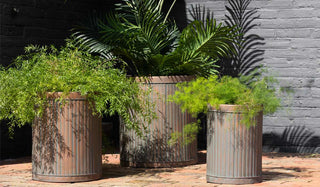 Lifestyle image of the Set Of 3 Antique Copper Garden Tub Planters displayed on a brick patio in the sunshine. 