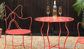 Red metal chair and table set with a bottle of wine and glasses on a gravel surface.