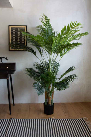 A very large, tall faux plant palm in a black pot, photographed by a desk by a striped rug. 