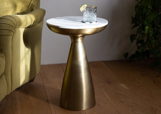 Image of a side table with a gold base and a round marble top. Photographed next to a green chair on a wooden floor. 