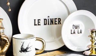 Lifestyle image of theSet Of 4 Le Diner Bistro Dinner Plates, Set Of 4 Ooh La La Bistro Bowls and Set of 2 Harry Tattoo Mugs styled together with other home accessories on a black sideboard. 