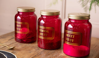 Three pink storage jars with gold lids and humorous labels on a wooden surface.