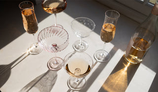 Various glasses with a wine bottle in the sunlight on a white surface.