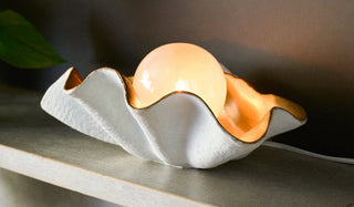 Lifestyle image of the Clam Table Lamp displayed illuminated on a fireplace shelf next to a plant.