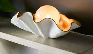 Lifestyle image of the Clam Table Lamp displayed illuminated on a fireplace shelf next to a plant.