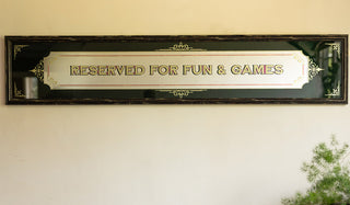 The Reserved For Fun & Games Vintage-Style Mirror displayed on a beige wall with a plant in the corner of the shot.