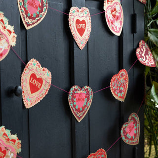 Lifestyle image of the Rockett St George Valentine Love Heart Paper Garland displayed across a black cabinet with greenery. 