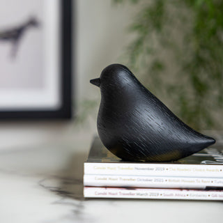 Lifestyle image of the Bobby The Black Bird Ornament displayed on a marble table on top of a stack of magazines.