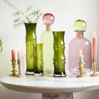The three sizes of the Green Glass Bamboo Vase, arranged with the Small and Large Pink & Green Apothecary Bottles and gold candlesticks with lit candles on a white marble table.