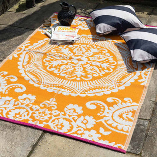 The Recycled Vintage Design Outdoor Rug in Orange styled outdoors in the sunshine with striped cushions, magazines, a jug and a glass.