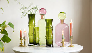 The Green Glass Bamboo Vases displayed with the Green & Pink Glass Apothecary Bottles with some gold candlesticks on a table next to a plant.