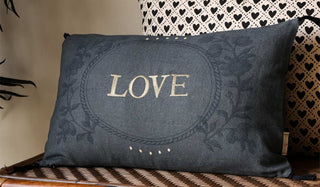 The Embroidered Love Cotton Cushion displayed on a wicker bench with another cushion, in front of a neutral wall with a plant to the left-hand side of the shot.