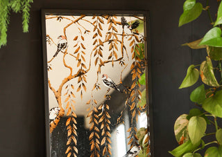 Lifestyle image of the Weeping Willow Tree & Bird Mirror displayed leaning against a black wall next to plants.