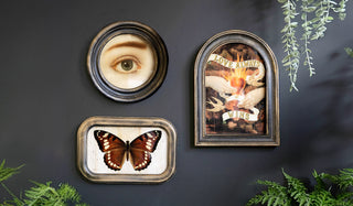 Three decorative framed pieces of wall art styled on a black wall surrounded by greenery.