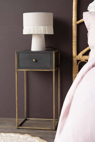 Lifestyle image of the entire Sophos Single Drawer Bedside Table next to a bed with a lamp on top with dark wooden flooring and dark wall background