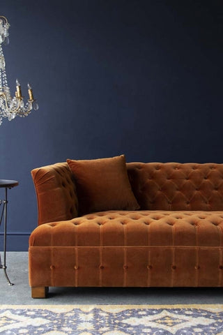 Lifestyle image of Burnt Orange Velvet Chesterfield Sofa with chandelier and rug on floor with dark wall background