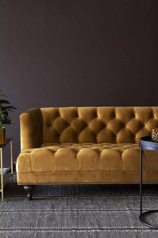 Lifestyle image of Ochre Gold Velvet Chesterfield Sofa with grey rug flooring and dark wall background