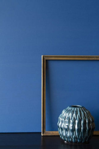 Blue 'Azurite' paint shown painted on a wall, displayed together with a wooden photo frame and textured blue vase. 