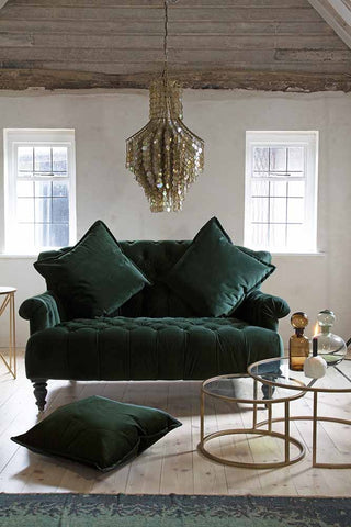 Lifestyle image of the Two-Seater Forest Green Velvet Sofa with cushions and Set Of 2 Circular Circus Nesting Tables and Shimmering Shell Disc Chandelier