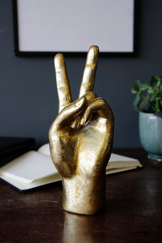 lifestyle image of gold peace hand ornament on wooden table with open book and plant